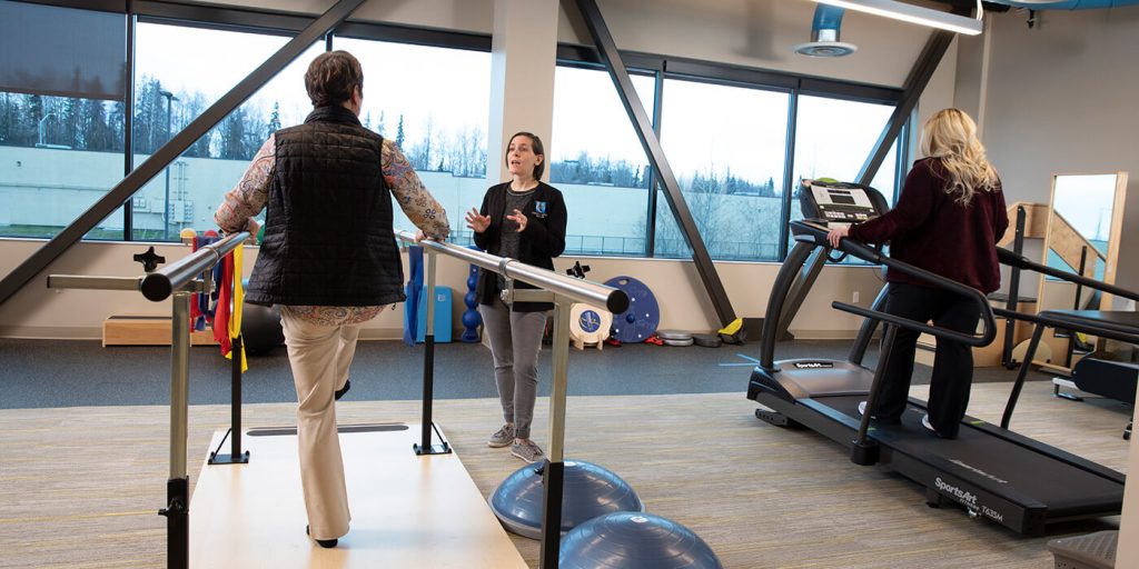 A woman talks to a doctor while standing on one foot in a physical therapy office.